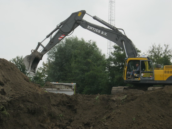 Een kundige kraanmachinist met zijn grijper op rupsbanden bezig de grote bulten af te graven. Afvoer met vrachtwagens... Wat wordt er een grond aangesleept en weer afgevoerd....