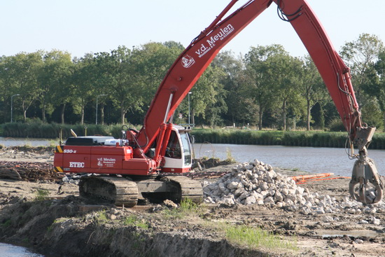 Doorlopen richting spoorbrug. Daar is een grijper gestaag bezig de brokken steen op de nieuwe oeverkant te deponeren.